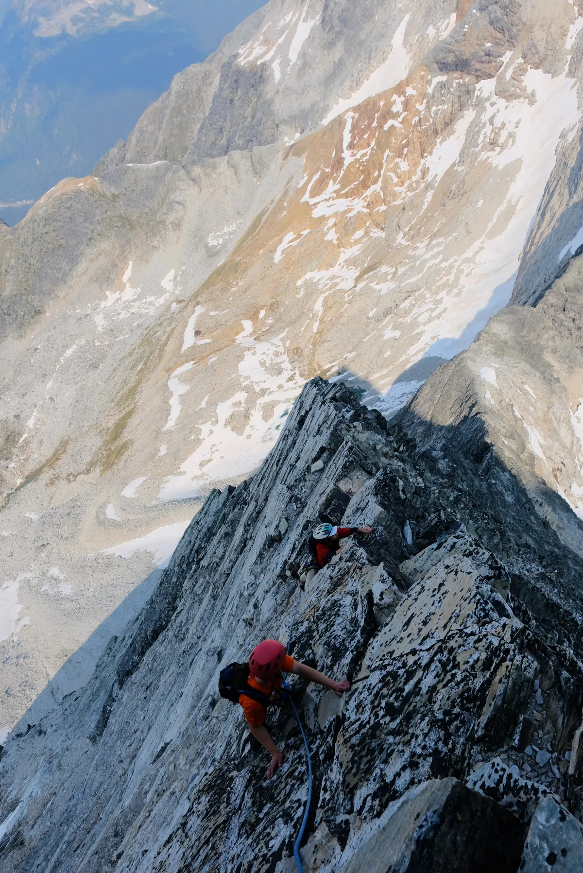 Looking down the ridge at two climbers following up the rock, scree and snow far below.