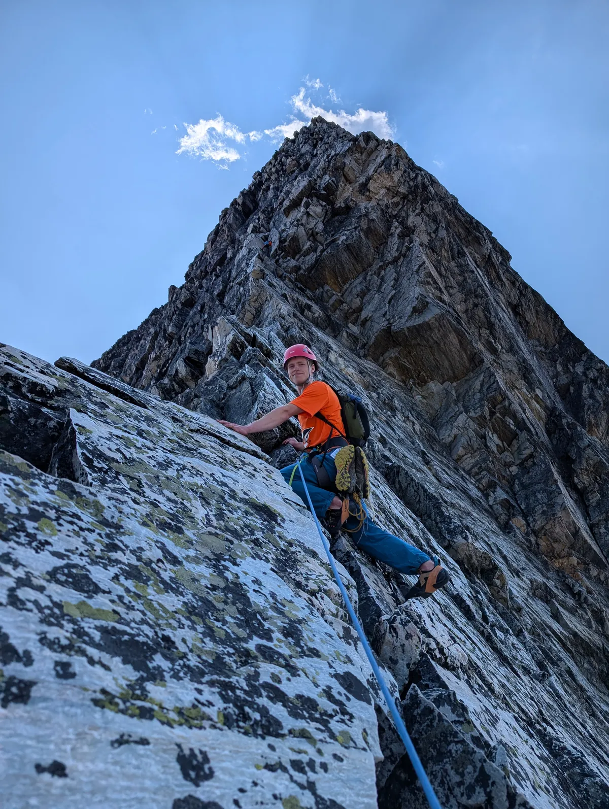 A climber on the slab looking back at the camera, rope trailing, the jagged summit above.
