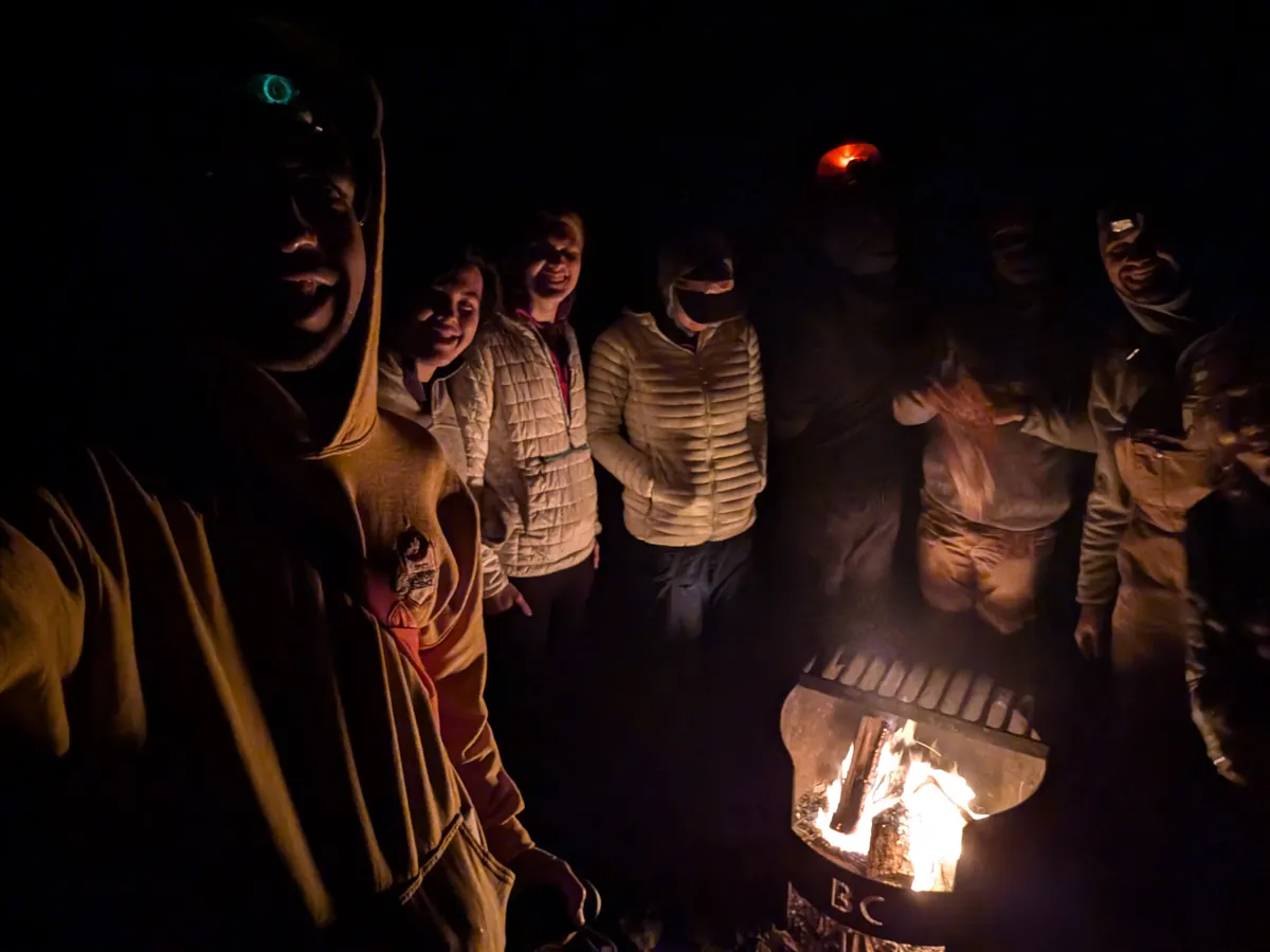 The group crowded around a fire pit in the dark, faces lit by the flames.