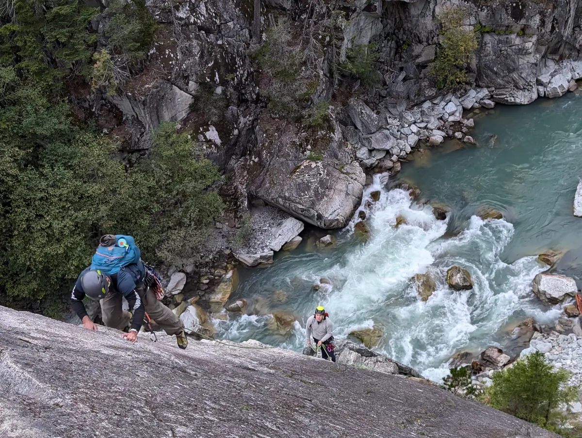 A climber with a large pack starting up the granite slab, whitewater rushing through the canyon below.