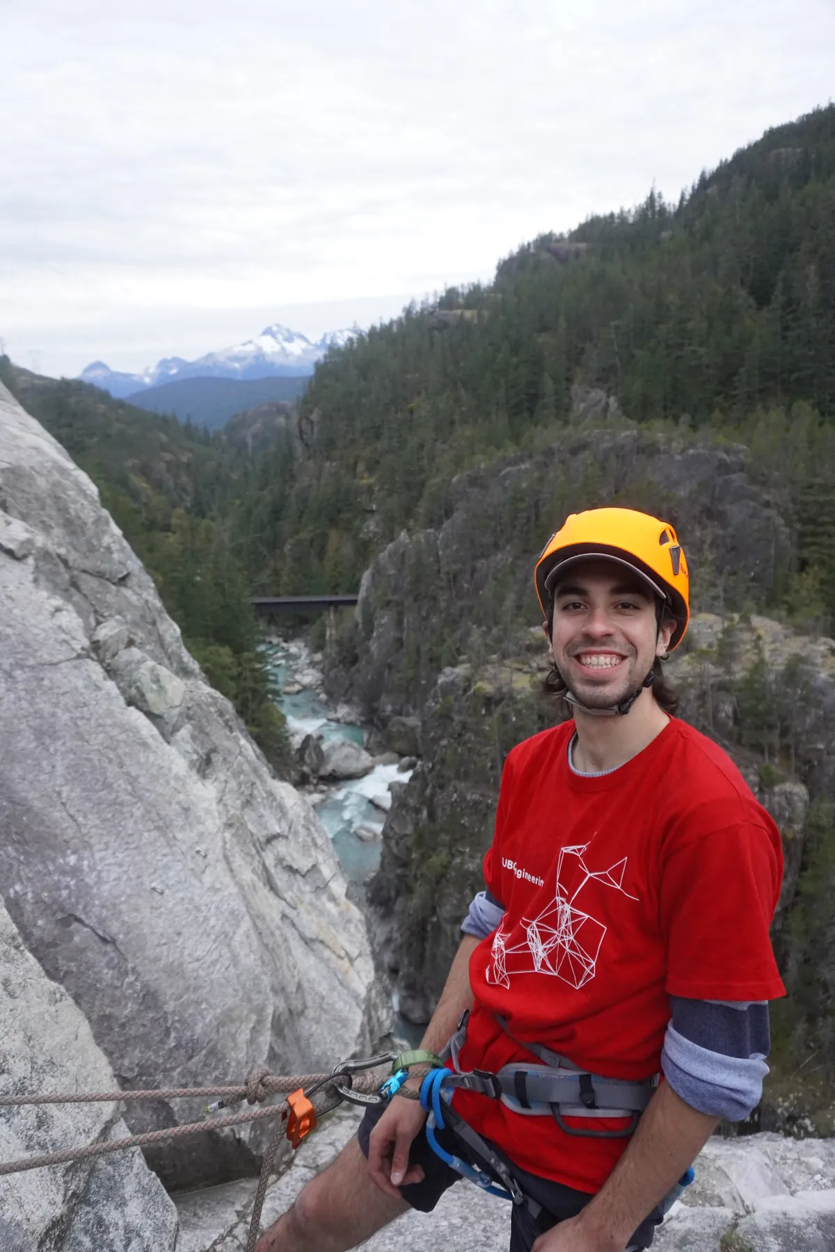 A climber grinning on a ledge in helmet and harness, the river gorge and distant snow-capped peaks behind.