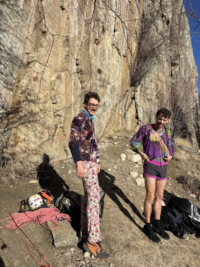 Two climbers in thrifted floral and purple outfits at the base of the wall, ropes and gear at their feet.