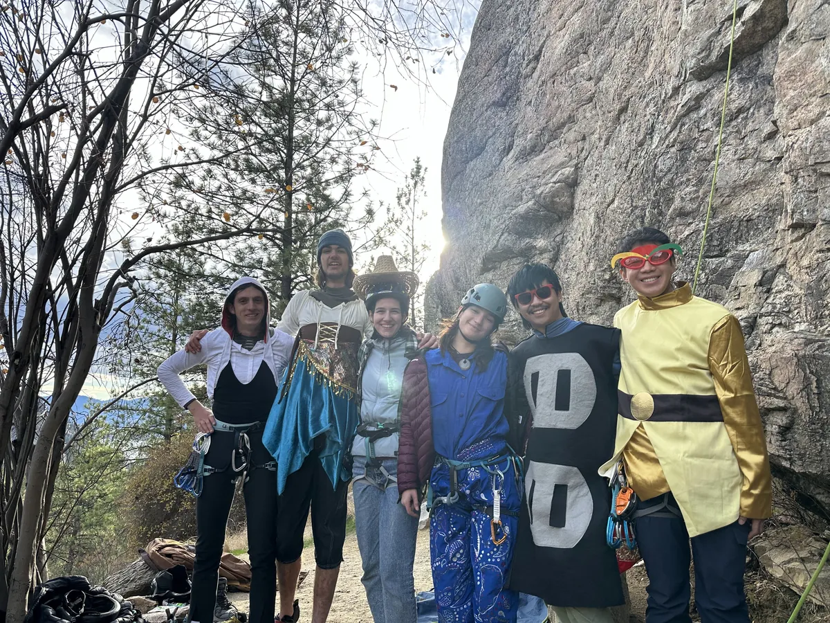 Six climbers in thrift-store costumes posing at the base of the crag, sun flaring through the trees.